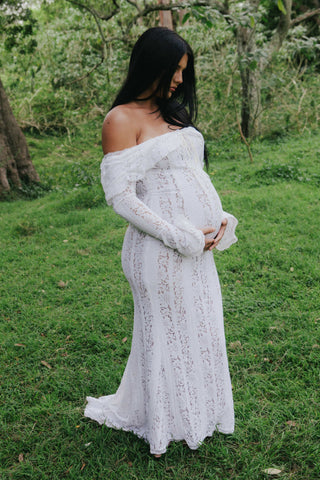A pregnant woman wearing the House of CB Belle Vintage Cream Floral Lace Off Shoulder Maxi Dress from Mama Rentals, standing gracefully on a grassy field surrounded by trees.