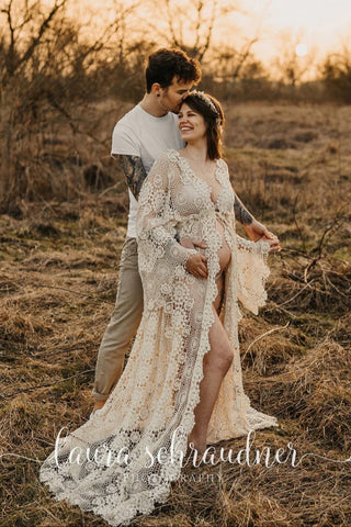 A pregnant woman wearing a beige crochet robe with long sleeves and a flowy design, standing in a field with a man behind her, both smiling during her maternity photoshoot. 