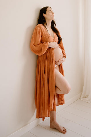 A pregnant woman posing in a warm rust-orange long-sleeve maternity robe  (Mama Rentals Jen's Pirate Booty Lapis Maxi Dress - Rust) against a minimalist off-white wall and light wood floor.