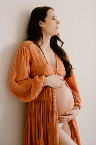 A pregnant woman wearing a textured rust-orange long-sleeve maternity robe (Mama Rentals Jen's Pirate Booty Lapis Maxi Dress - Rust), leaning against a plain off-white wall.