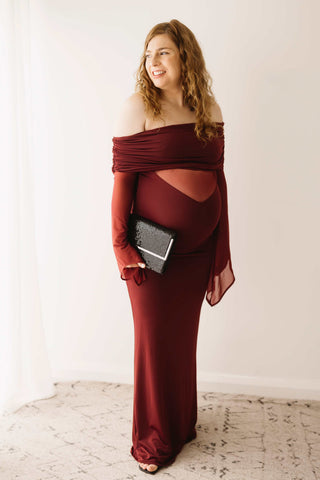 A pregnant lady wearing the Mama Rentals Meshki Kaitlin One Shoulder Slinky And Mesh Maxi Dress - Burgundy holding the Olga Berg Black Sequin Clutch in a studio.