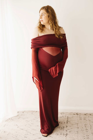 A pregnant lady wearing the Mama Rentals Meshki Kaitlin One Shoulder Slinky And Mesh Maxi Dress - Burgundy in a studio.