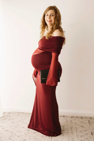 A pregnant lady wearing the Mama Rentals Meshki Kaitlin One Shoulder Slinky And Mesh Maxi Dress - Burgundy holding the Olga Berg Black Sequin Clutch in a studio.