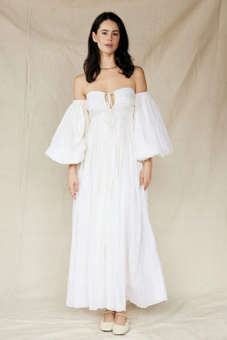 Woman wearing a Rooh Collective Bronte Maxi Dress - a white off-shoulder dress with bell sleeves against a beige background