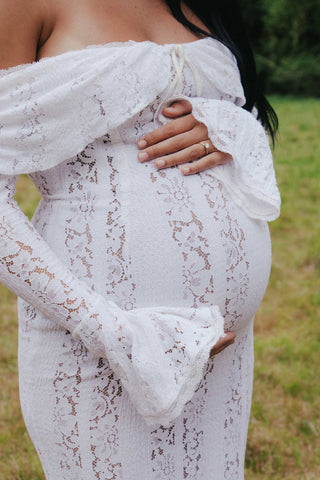 Pregnant woman embracing her belly in Mama Rentals’ House of CB Belle Vintage Cream Floral Lace Off Shoulder Maxi Dress, surrounded by soft greenery and trees.