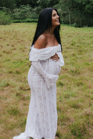 Mama Rentals’ House of CB Belle Vintage Cream Floral Lace Off Shoulder Maxi Dress styled by an expecting mother in a serene outdoor setting, gently holding her baby bump.