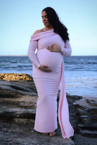 A pregnant woman stands on a rocky beach wearing a light pink off-the-shoulder long-sleeve maternity gown with a side slit and a long draped train, the Mama Rentals Club L London Amara Maternity Maxi with Sash.
