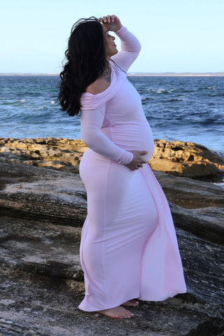 A pregnant woman wearing a light pink off-the-shoulder maternity maxi dress, the Mama Rentals Club L London Amara Maternity Maxi with Sash, standing barefoot on a rocky shoreline with the ocean and sky in the background.