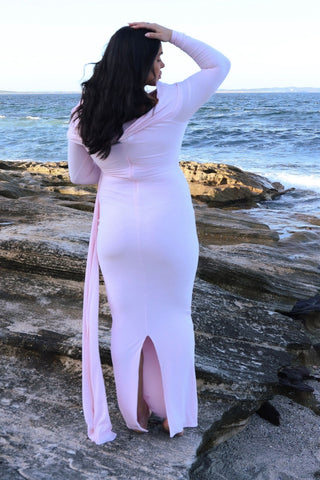 A woman stands on rocky shoreline facing the ocean, wearing a fitted long-sleeve maxi dress in a very pale pink with a side slit and a long draped tie detail at the back, the Mama Rentals Club L London Amara Maternity Maxi with Sash.