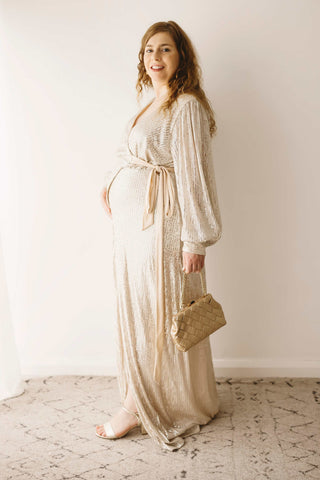A pregnant woman wearing the Mama Rentals Jasmine Sequin Gown - Champagne holding the Olga Berg Quinn Woven Gold Clutch in a studio.
