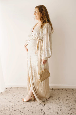 A pregnant woman wearing the Mama Rentals Jasmine Sequin Gown - Champagne holding the Olga Berg Quinn Woven Gold Clutch in a studio.