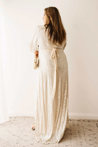 A woman showing the back design of the Mama Rentals Jasmine Sequin Gown - Champagne holding the Olga Berg Quinn Woven Gold Clutch in a studio.